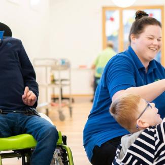 ein Kind im Rollstuhl das lacht, ein Kind kniend das mit einem Spilezug an der Wand spielt und eine Pflegerin die sich mit beiden beschäftigt