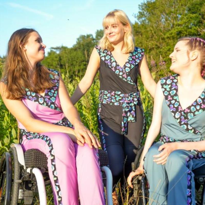 3 Frauen, zwei davon im Rollstuhl sitzend, tragen schön gestylte Overals und Hosenröcke