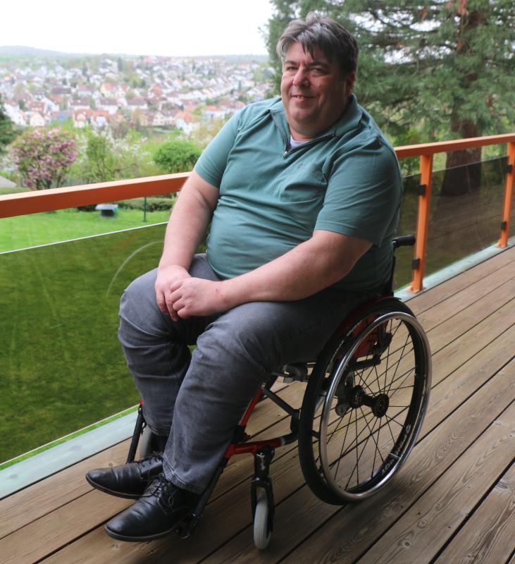 Bild zeigt Rollstuhlfahrer (dicklich) Alexander Lang auf seinem Balkon, im Hintergrund sieht man die Landschaft
