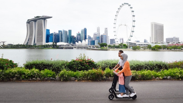 Bild zeigt 2 Erwachsene und ein Kind das zwischen den Beinen der Mutter auf einem E-Scouter steht, im Hintergrund dei Skyline von Singapor