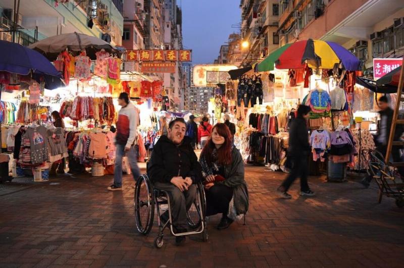Antony im Rollstuhl mit Begleitung kniend auf einem Markt in China