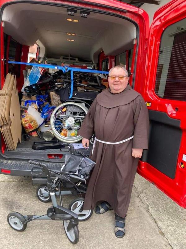 Franziskaner-Mönch in Ordenstracht steht lächelnd vor dem geöffneten Heck eines Kleintransporters. Zu sehen sind ein Teil der Hilfsmittel, wie ein Rollstuhl und Rollatoren.