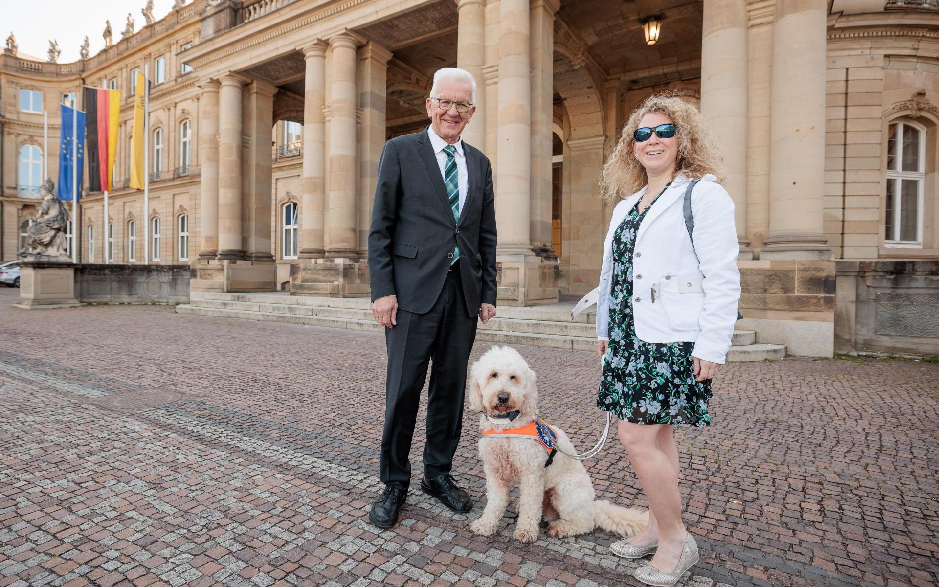 Winfried Kretschmann, der Ministerpräsident von Baden-Württemberg, steht links im Bild. Er lächelt in die Kamera. Er ist Mitte siebzig, hat weiße Haare, trägt eine Brille und einen schwarzen Anzug mit Krawatte. Rechts neben ihm sitzt der Hund Harry, ein Goldendoodle mit blondem, lockigem Fell. Er trägt eine Kenndecke und wird gehalten von Lisa Mümmler, rechts neben ihm. Sie ist eine junge Frau mit blondgelocktem Haar. Sie trägt eine schwarze Sonnenbrille, ein geblümtes Kleid und eine weiße Jacke. Sie lächel