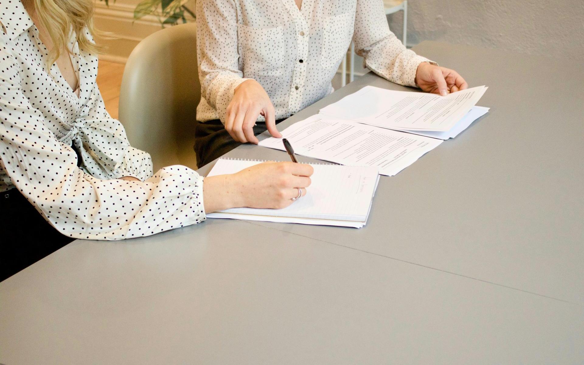 Bild zeigt einen Schreibtisch mit Papier. 2 Damen nur der Oberkörper ist zu sehen bearbeiten dieses Papier