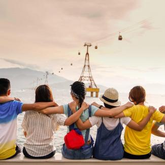 Gruppe mit jungen Menschen schaut arm in arm auf das Meer, oben links eine Seilbahn