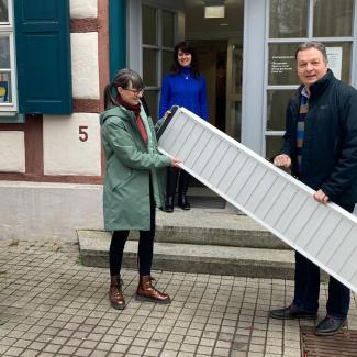 ein Mann übergiebt eine Rollstuhlrampe an einen Frau vor einem Museum in einem Fachwerkhaus. Der Eingang hat zei Stufen, in der Tür wartet eine weitere Frau